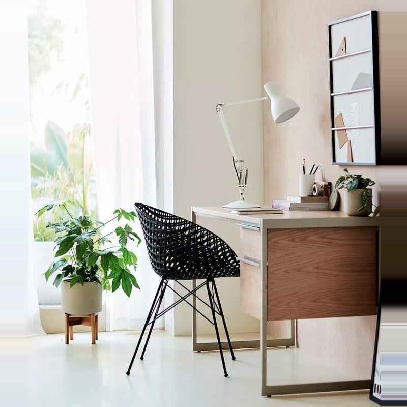 A stylish and minimalist workspace featuring a wooden desk with a modern white lamp, a black woven chair, and indoor plants, bathed in natural light from a large window with sheer curtains.