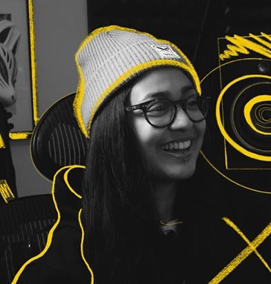 Portrait of a music producer wearing a gray beanie, glasses, and a hoodie, smiling in a creative studio environment with vibrant yellow graphic accents highlighting musical equipment, representing users of the Chillblast Spark Creator PC range.