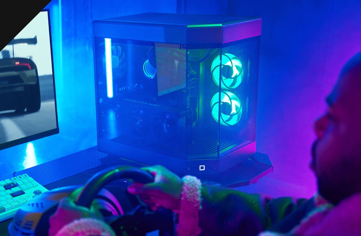 Chillblast Vantage PC with a compact, glass-panelled design and vibrant RGB-lit cooling fans, set up in a racing simulation environment, featuring a gamer using a steering wheel controller and a monitor displaying a high-speed racing game.