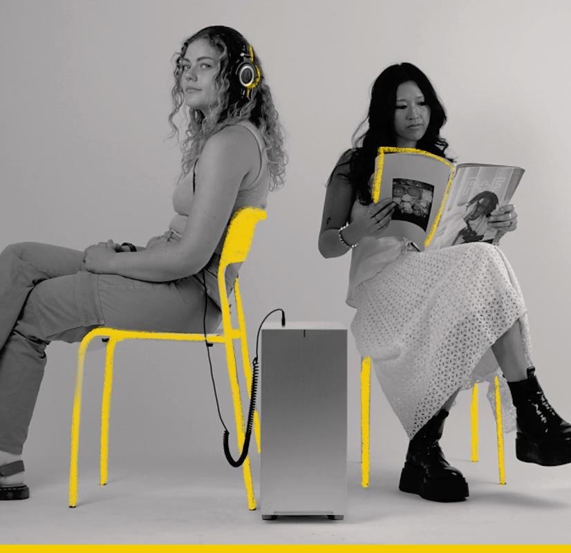 A Chillblast Spark Video Ultra Workstation computer positioned between two individuals seated on yellow-highlighted chairs; one wearing headphones and holding a controller, the other reading a magazine, symbolizing diverse creative uses of the workstation in a modern and minimalist setup.