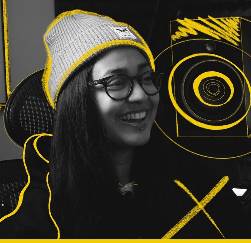 Portrait of a music producer wearing a gray beanie, glasses, and a hoodie, smiling in a creative studio environment with vibrant yellow graphic accents highlighting musical equipment, representing users of the Chillblast Spark Creator PC range.