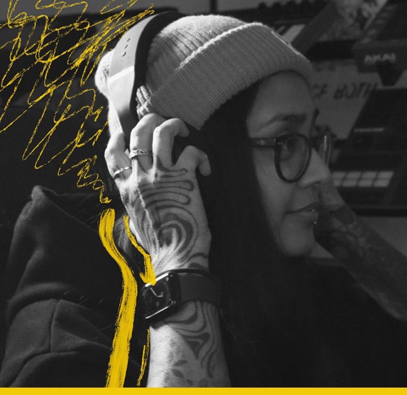 Close-up of a music producer wearing headphones and a beanie, with intricate tattoos on their hand and vibrant yellow highlights accentuating the creative atmosphere, surrounded by music production equipment.