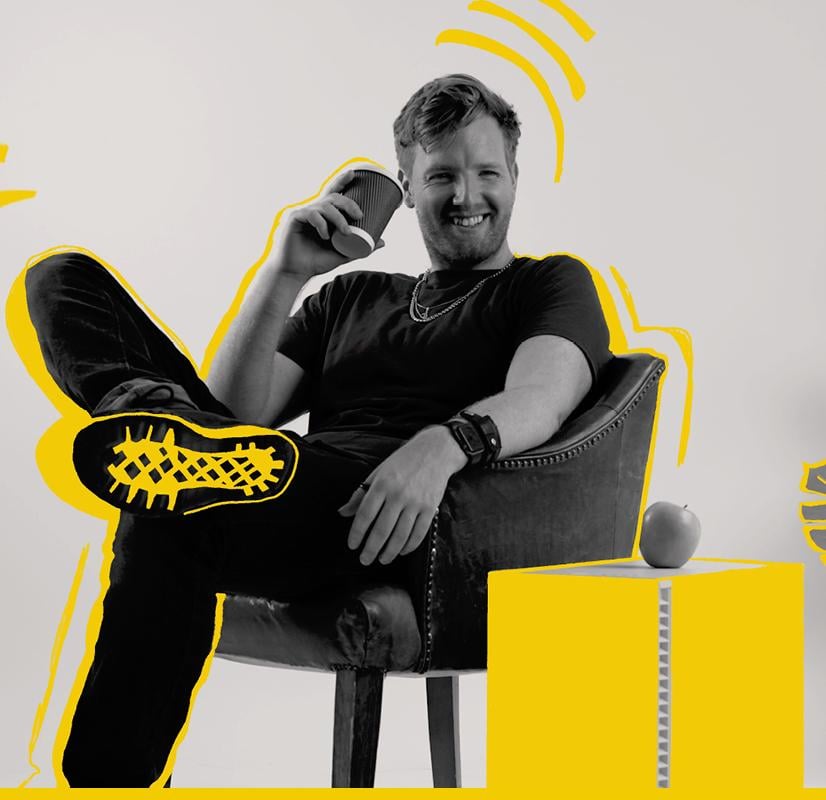 Portrait of a photographer lounging in a chair, holding a coffee cup and smiling, with bold yellow highlights emphasizing their pose and surroundings, including a Chillblast Spark Video Ultra Workstation and an apple, symbolizing creativity and relaxation.