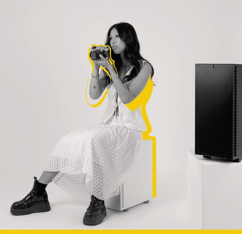 A photographer seated on a Chillblast Spark Video Ultra Workstation, holding a vintage camera and dressed in a stylish white outfit, with bold yellow highlights accentuating the composition, symbolizing creativity and artistic expression.
