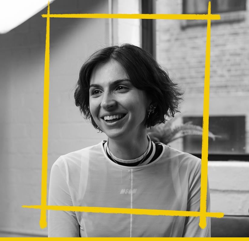 Portrait of a 3D designer with a short haircut and a bright smile, wearing a light sheer top, set in a modern workspace with yellow graphic accents framing the image, representing the creative professionals who use the Chillblast Spark Creator PC range.